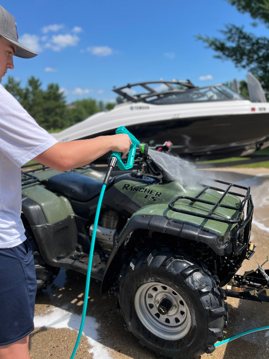 A person in a white t-shirt and navy shorts washes a green and black Ranger XSV ATV with a turquoise hose nozzle, with a white and black boat parked in the background under a blue sky.