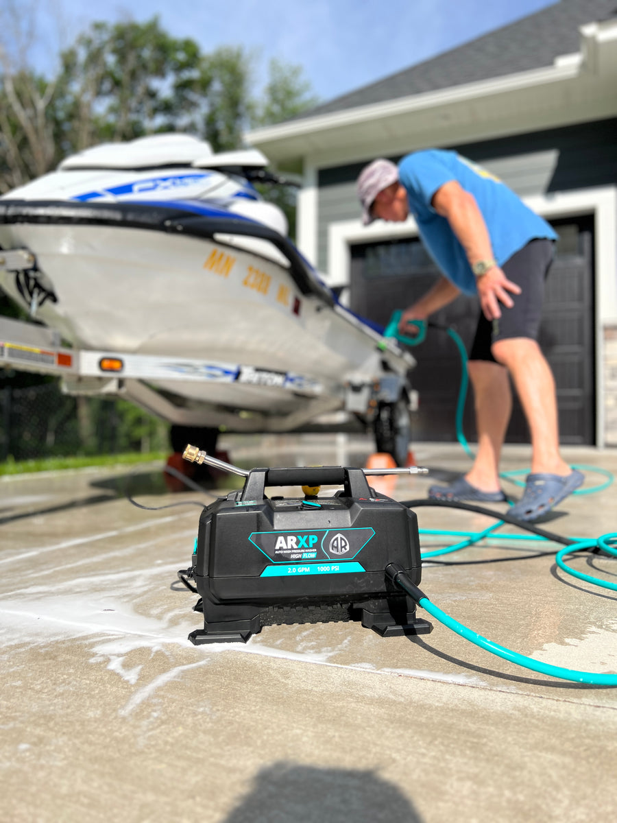 A black ARXP pressure washer sits on a concrete driveway in front of a boat trailer and a man washing a boat. The ARXP unit is connected to a bright green hose.
