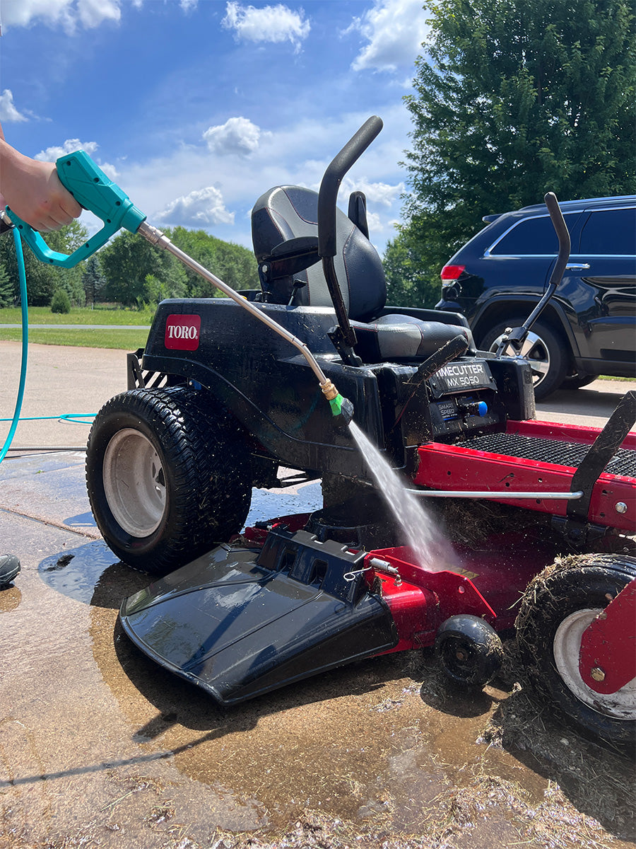 A red and black riding lawn mower being washed with a hose outdoors on a wet concrete surface, with a person's hand holding the hose and a blue sky with trees in the background.